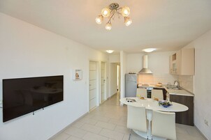 Cozinha moderna com televisão de tela plana, área de refeições com mesa e cadeiras brancas, e corredor que leva a outro quarto.