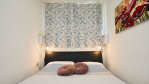 Un letto rifatto con due cuscini rotondi, un lampadario a parete, un'opera d'arte a muro e una finestra con tenda a motivi foglia.