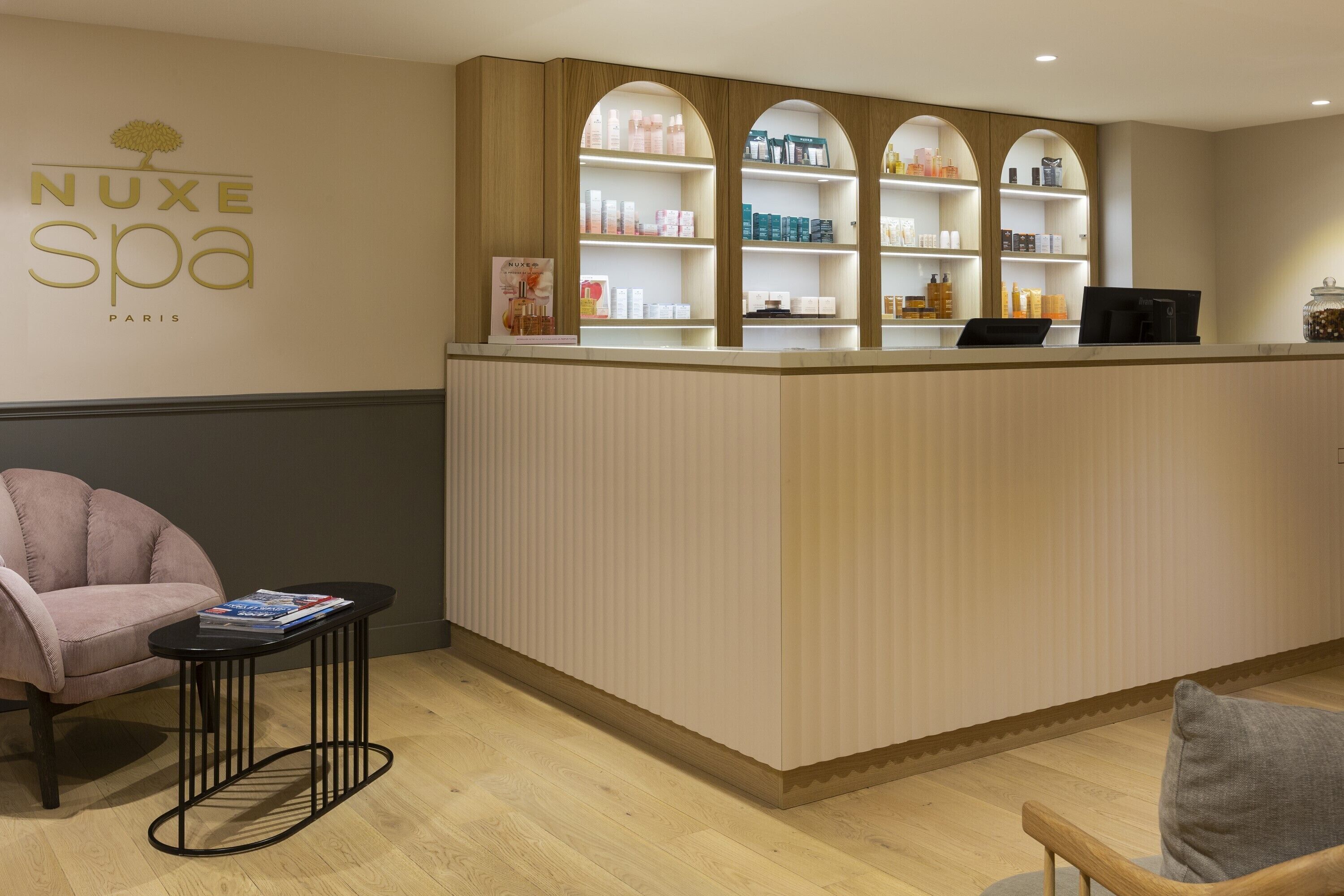 Un espace d’accueil comprenant un comptoir de réception, une chaise et une étagère où sont présentés des produits de spa.