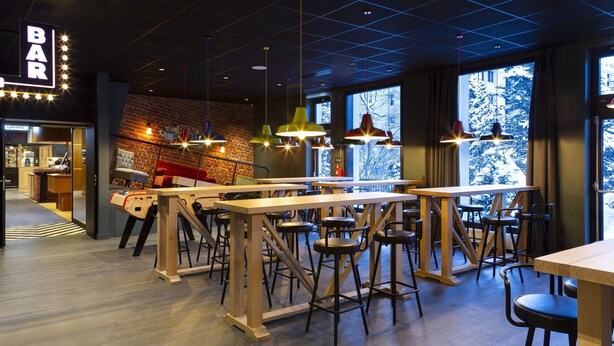 Un espace bar moderne avec des tables en bois et des tabourets de bar, de grandes fenêtres offrant une vue sur le paysage enneigé, et un mur de briques agrémenté d’une enseigne au néon.