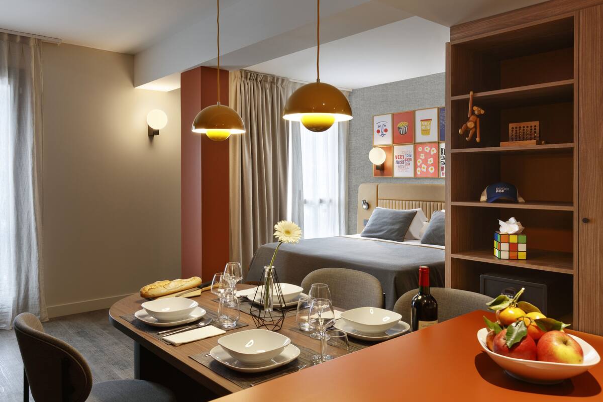 Une chambre d’hôtel bien rangée, avec une table à manger dressée pour deux personnes, un lit recouvert d’une couverture grise, une table de chevet avec une bouteille de vin et une étagère contenant divers objets, dont un Rubik’s Cube.