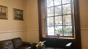 A room with a brown leather sofa, a wooden coffee table with a vase of flowers, and a large window with a view of buildings and a tree. - Europa House (London)