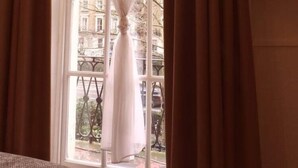 A room with a window featuring a white curtain tied at the top, a view of a cityscape with buildings and trees, and a brown curtain on the left side. - Europa House (London)