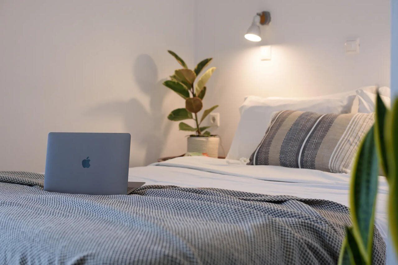 Un letto rifatto con cura, un laptop sopra, una pianta sul comodino e una lampada a parete.