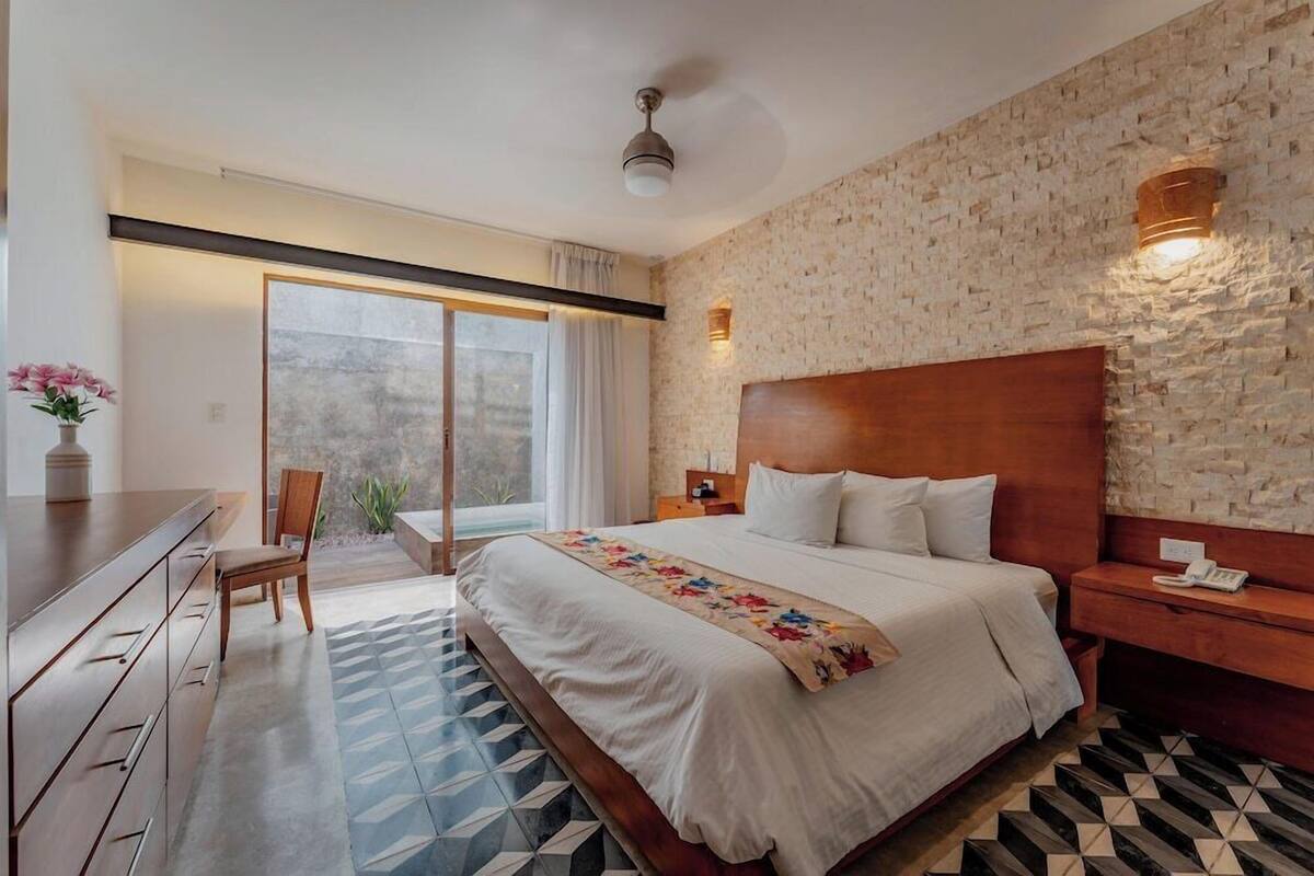 Ein Hotelzimmer mit einem großen Bett, einem Nachttisch aus Holz, einem Schreibtisch mit Schubladen, einem Sessel und einem großen Fenster mit Vorhängen, sowie einer Steinwand.