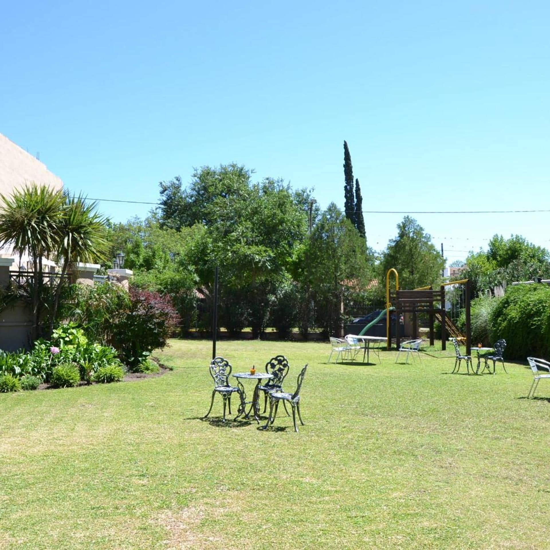 Un espacio al aire libre con césped, una mesa y sillas, rodeado de árboles y un parque infantil.