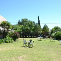 Un espacio al aire libre con césped, una mesa y sillas, rodeado de árboles y un parque infantil.