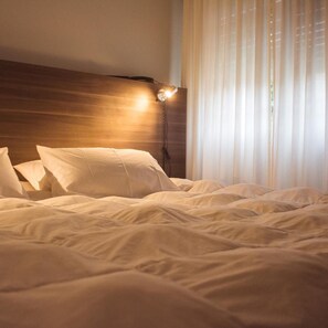Un lit bien fait, avec du linge de lit blanc et des oreillers, une tête de lit en bois et une lampe fixée au mur.