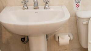Une salle de bain avec un lavabo blanc, deux robinets, des toilettes et un rouleau de papier toilette.