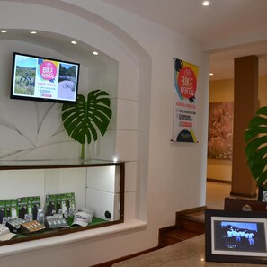 A reception area with a TV screen displaying a bike rental advertisement, a large plant, and a framed photo on a counter. - Solares Del Alto Hotel And Spa (Alta Gracia)