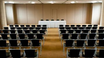 Sala de conferências com fileiras de cadeiras voltadas para uma mesa branca, paredes bege e piso coberto por carpete.