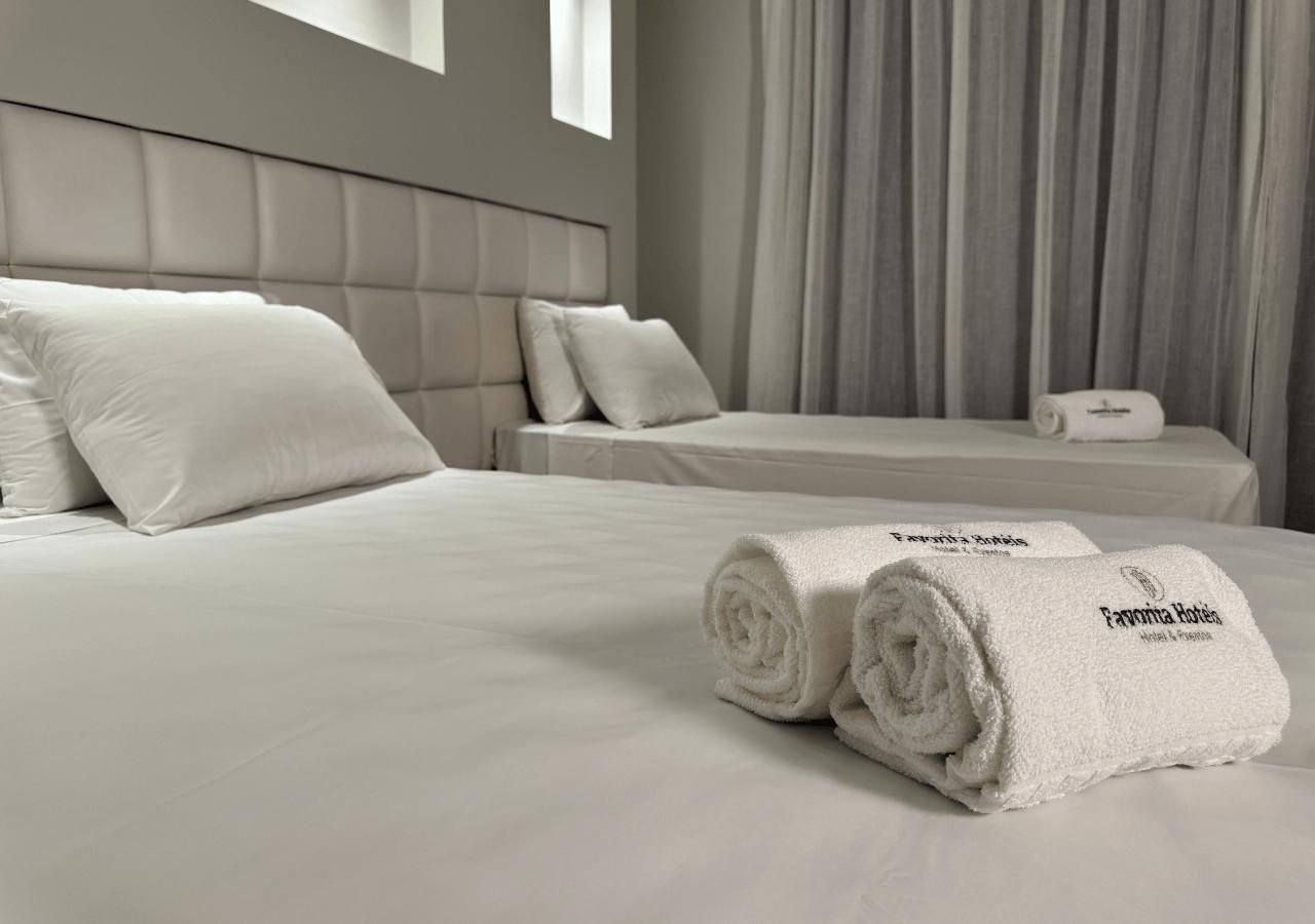 A hotel room with a large bed, two pillows, a rolled towel, and a bedside table with a bottle.