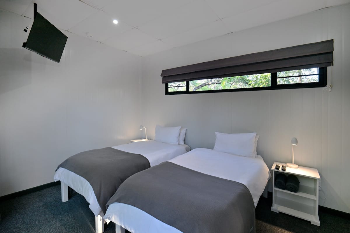 Habitación de hotel con dos camas, una ventana con vistas, un televisor montado en la pared y una mesita de noche con lámpara.