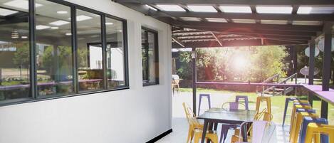 A covered outdoor dining area with tables and chairs, large windows, and a view of greenery.