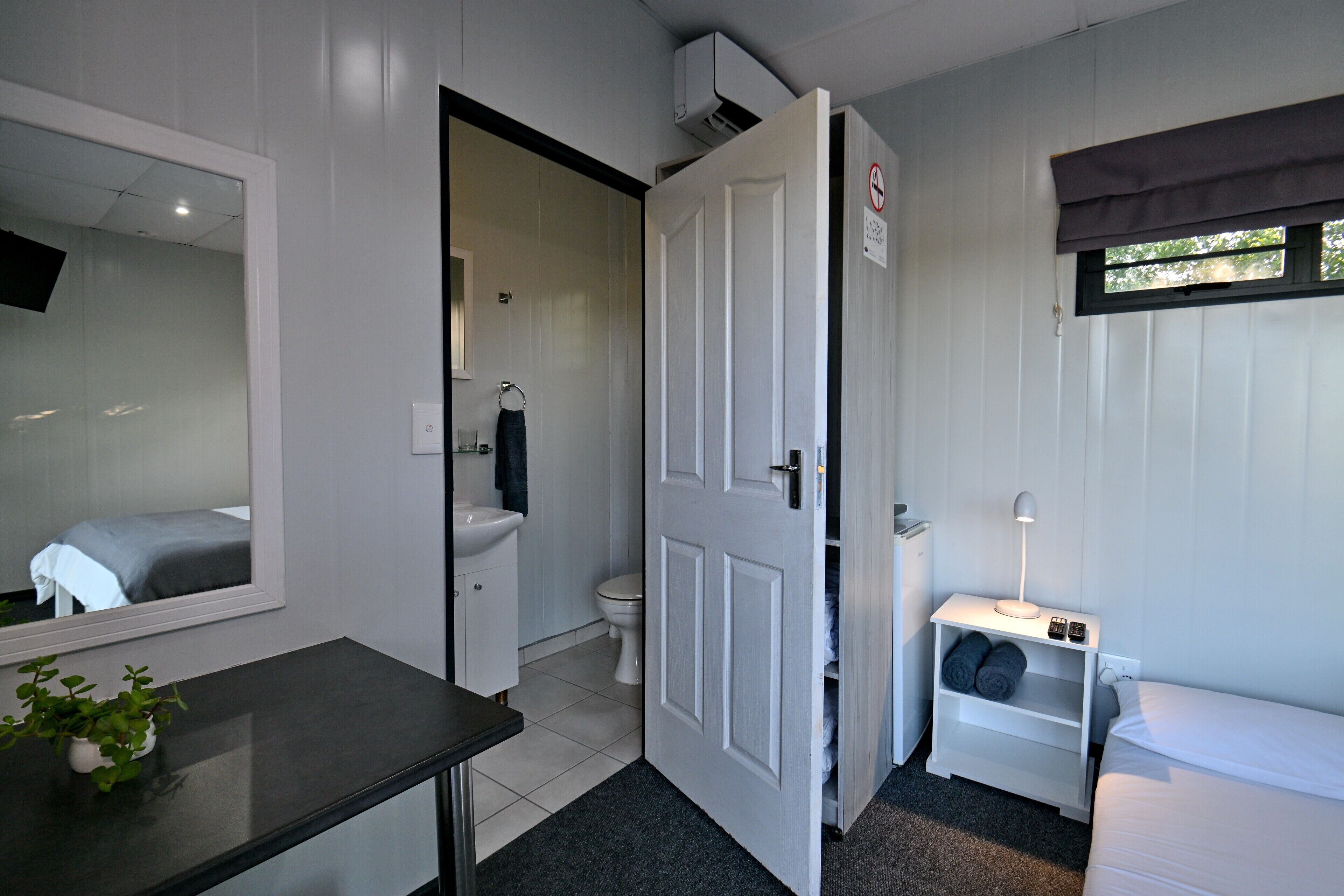 A hotel room with a bed, a bedside table, a nightstand, a mirror, a toilet, and a sink.