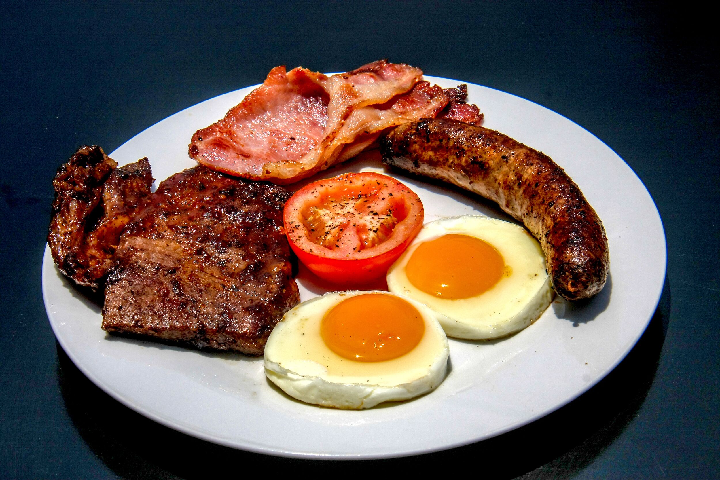 A plate with bacon, sausage, steak, two eggs, and a tomato slice.