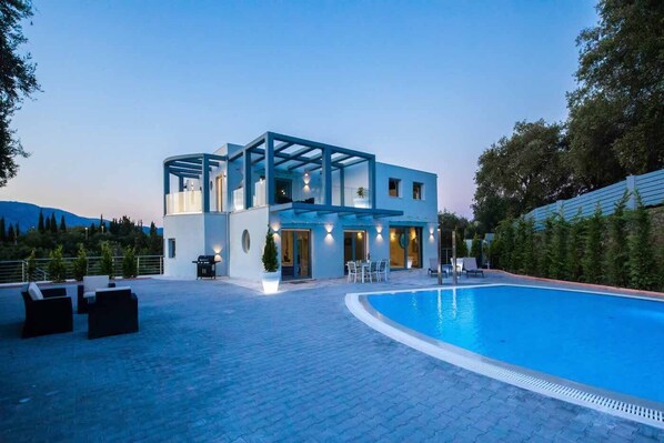 A modern two-story building with a swimming pool, outdoor seating, and a view of the mountains.