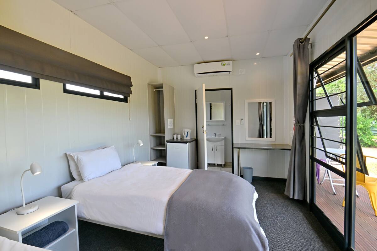 A modern hotel room with a bed, bedside table, desk, and a view of the outdoors through a large window.