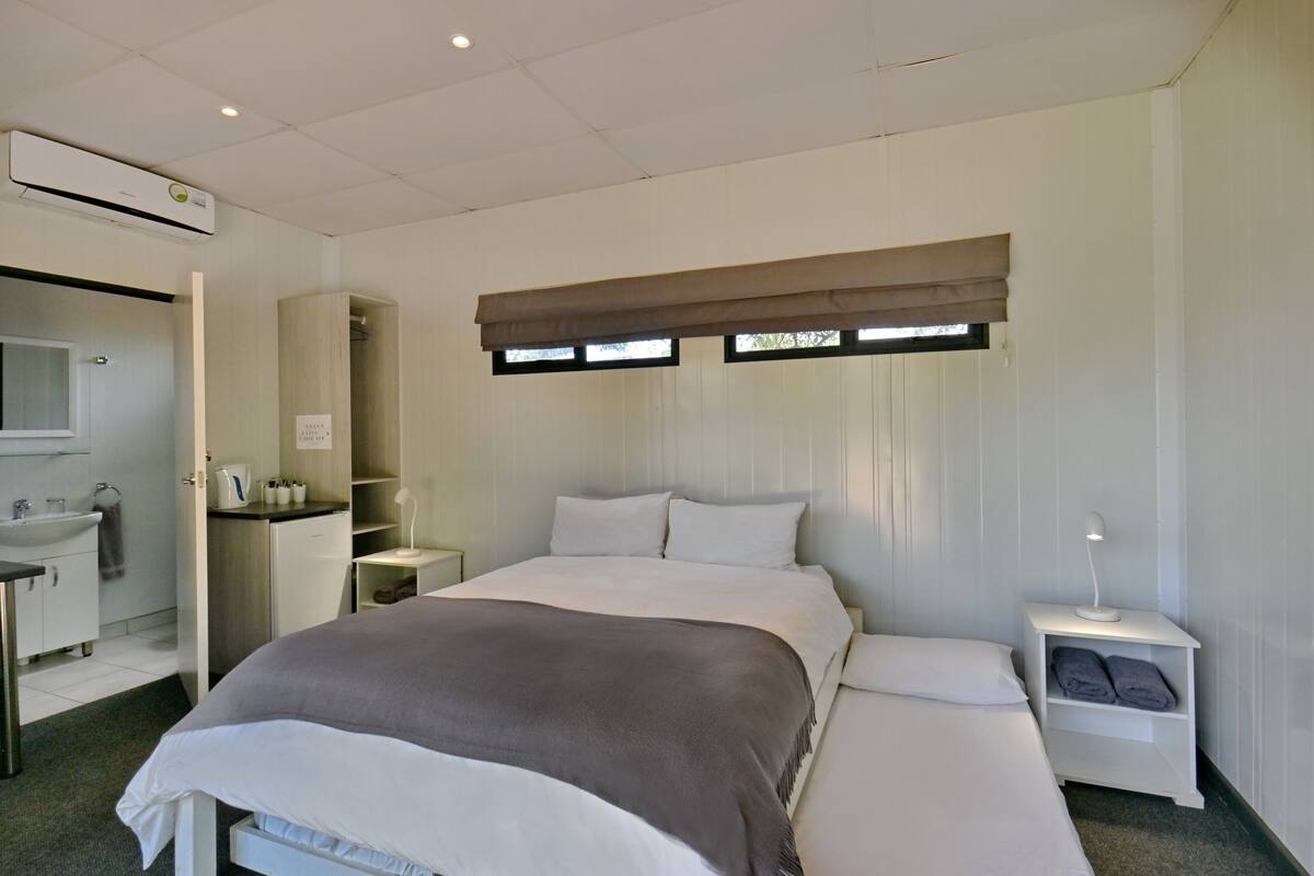A hotel room with a bed, bedside table, lamp, and a window with blinds.