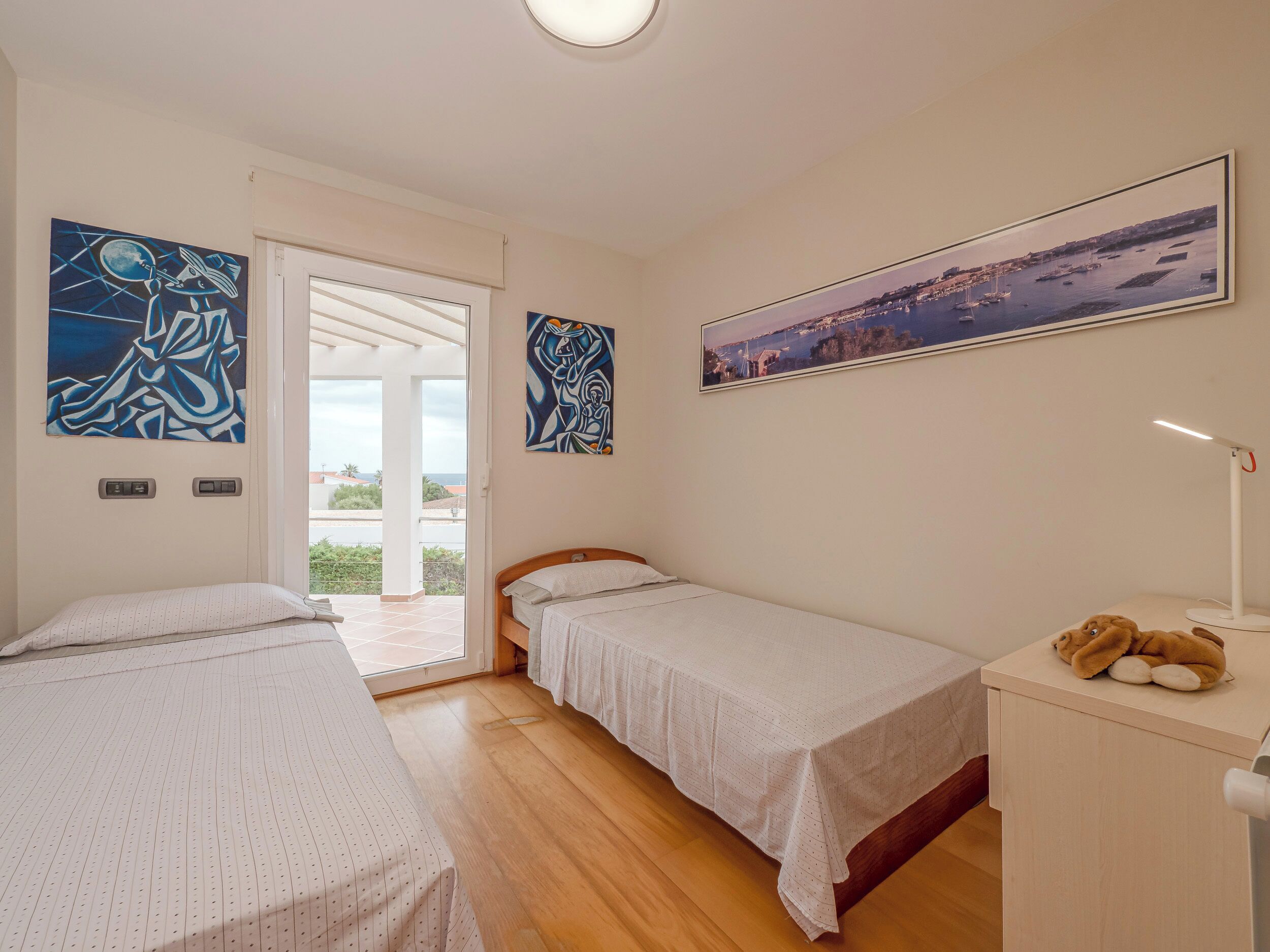 A bedroom with two single beds, a wooden floor, a desk with a lamp, and two paintings on the wall.
