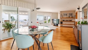 Een moderne woonkamer met een glazen eettafel, twee lichtblauwe stoelen, een bank, een televisie en een keukenhoek met een koelkast en planken.