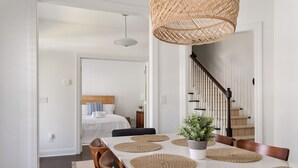 A dining area with a white table, brown chairs, and woven placemats. A potted plant is on the table, and a wicker chandelier hangs above.