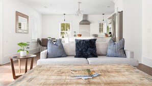 Una sala de estar moderna con un sofá gris claro, dos almohadones estampados, una mesa de centro redonda de madera con revistas, una mesita redonda con una planta en maceta, y una cocina al fondo.