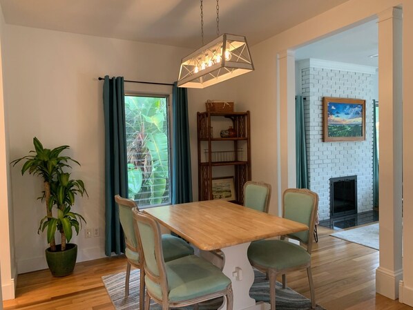 Un coin repas avec une table en bois et quatre chaises, un luminaire suspendu, une bibliothèque, une plante en pot et une vue sur un mur de briques et une cheminée.