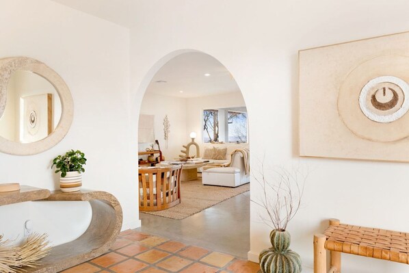 A modern interior with a curved stone sink, a round mirror, a wooden bench, and a living area with a sofa and coffee table.
