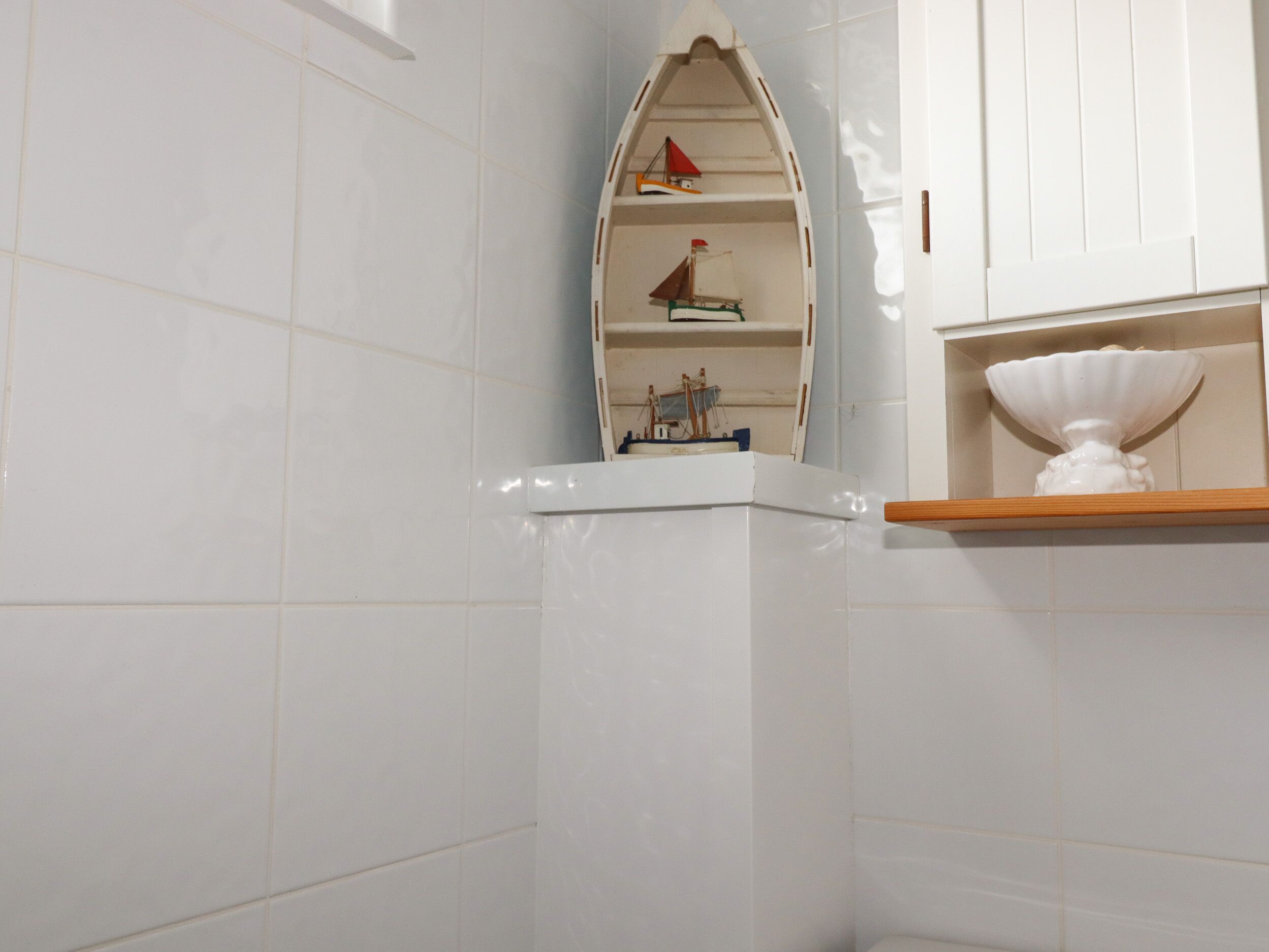 Une salle de bain aux murs recouverts de carreaux blancs, une étagère d’angle ornée d’un petit modèle de bateau et un bol en céramique blanche posé sur une étagère en bois.