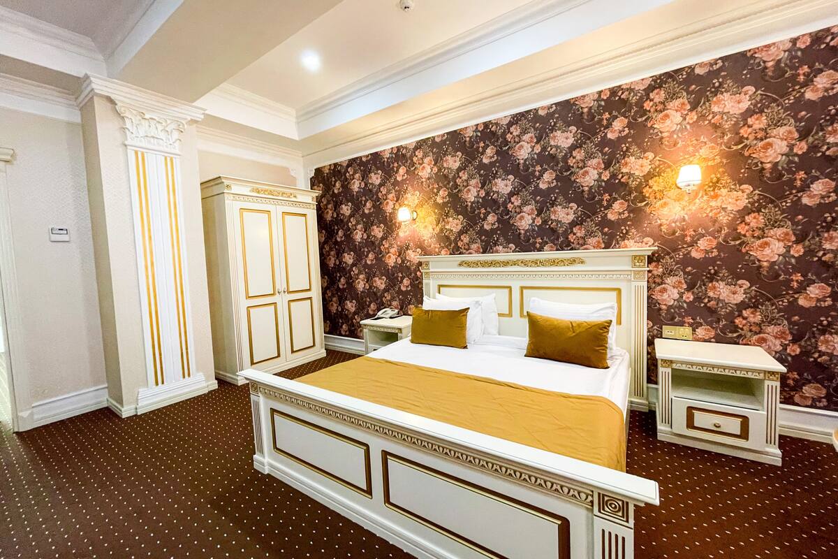 A bedroom with a large bed, two bedside tables, a wardrobe, floral wallpaper, and a patterned carpet.