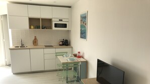 Cocina moderna con armarios blancos, microondas y lavabo. Hay un comedor con mesa y sillas, y un televisor de pantalla plana sobre un soporte de madera.