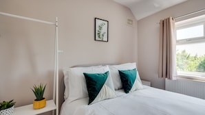 Ein Schlafzimmer mit einem Bett, einem Nachttisch, einer Topfpflanze, einem gerahmten Bild und einem Fenster mit Vorhängen.