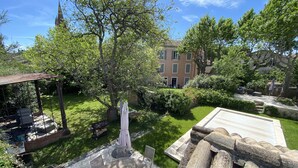 A courtyard with a wooden table and chairs, surrounded by lush greenery and a building with a tower in the background. - Town house with swimming pool and private garden (Pélissanne)