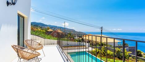 A balcony with a pool, two wicker chairs, and a view of the sea and hills.