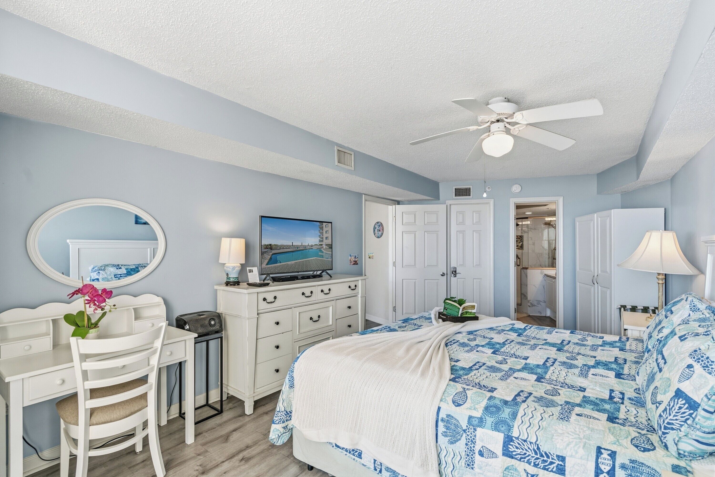 A bedroom with a bed, a desk, a chair, a television, and a ceiling fan.