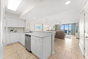 A modern kitchen with white cabinetry, a dishwasher, and a large island. There is a dining area with a sofa and a view of the outdoors.
