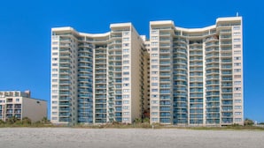 Un hotel de gran altura con múltiples balcones, un techo plano y una playa arenosa al frente.