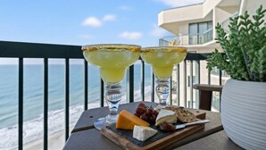 Un balcon avec vue sur l’océan, deux margaritas, du fromage, des raisins et des crackers disposés sur un plateau.