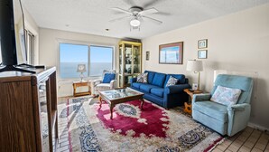 A living room with a ceiling fan, a blue sofa, a red and white patterned rug, a glass coffee table, and a blue armchair.