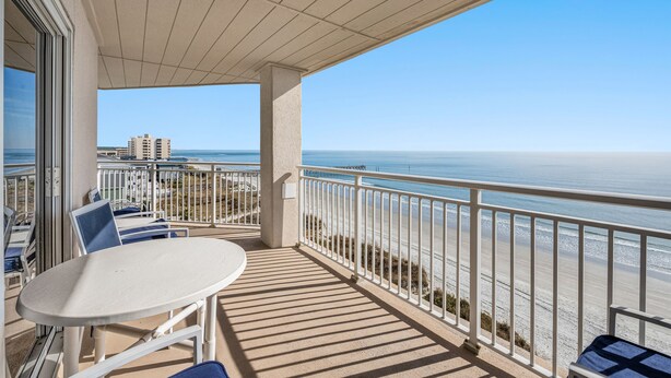 A balcony with a table and chairs, overlooking a beach with a clear view of the ocean and distant buildings.