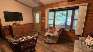 A cozy cabin-style room with wooden walls, a large window with curtains, a television mounted on the wall, and two upholstered chairs with patterned throw pillows.