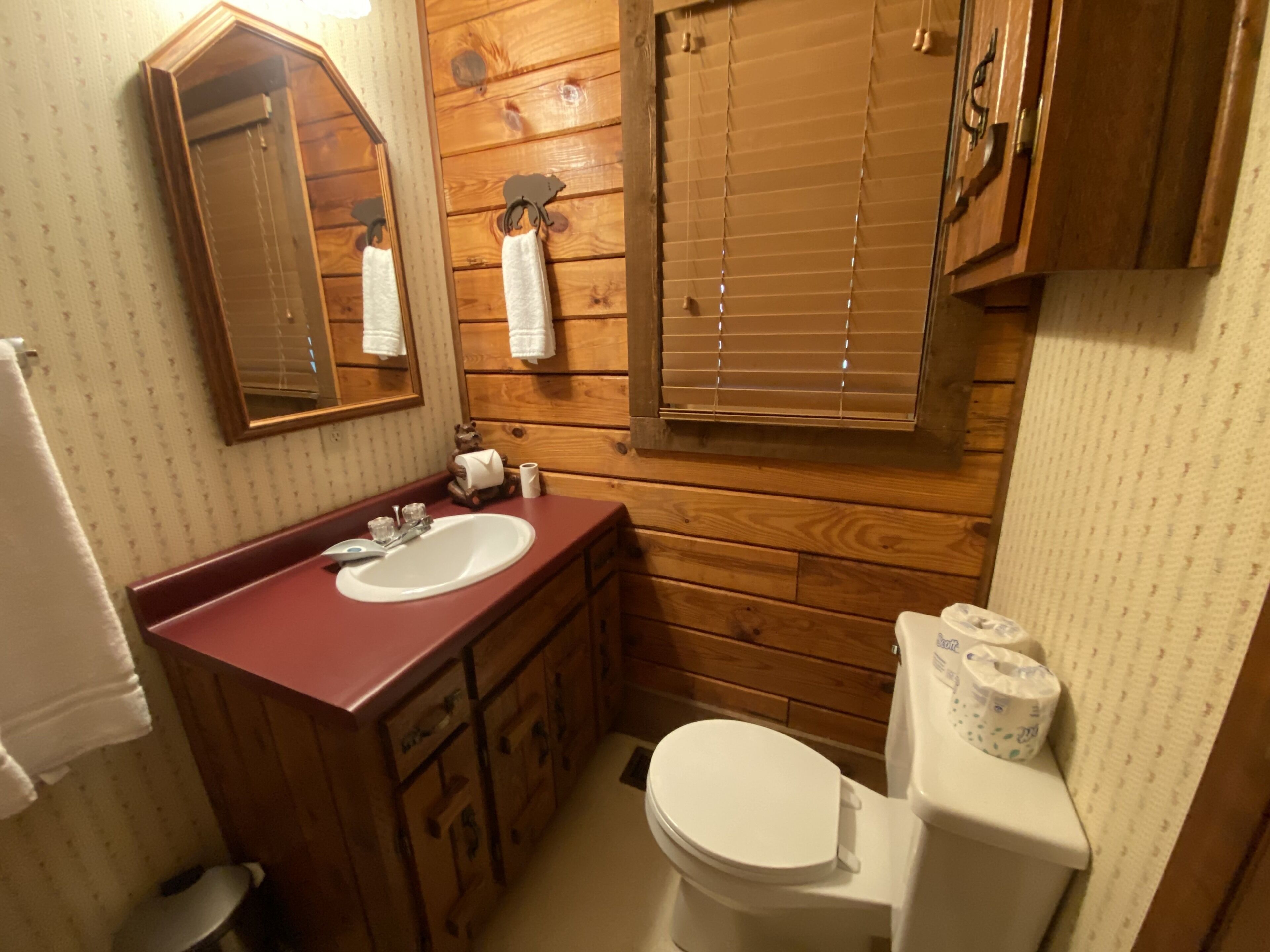 Un baño con paredes de madera, un lavabo, un espejo, un inodoro y una ventana con persianas.