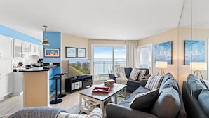 A modern living room with a sofa, a coffee table, and a kitchen area with a bar stool. There are framed pictures on the wall and a view of the city through the window. - Xanadu II 605-3BR/3BA Oceanfront condo in Cherry Grove (North Myrtle Beach)