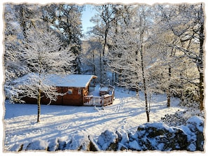 A wooden cabin with a deck, surrounded by snow-covered trees and a clear sky. - Blelham Tarn (Rustic yet modern cabin in tranquil woodland with own EV charger) (Ambleside)
