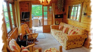 A cozy cabin interior with a sofa, chairs, a television, and a wooden deck visible through the door. - Blelham Tarn (Rustic yet modern cabin in tranquil woodland with own EV charger) (Ambleside)