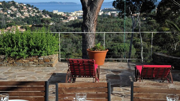 An outdoor dining area with stone flooring, wooden chairs, and a table set for two. A large potted plant is in the center, and the background features a scenic view of a coastal town. - Beautiful villa with sea view, 2.5km from the beach, in the heart of nature   (Bormes-les-Mimosas)
