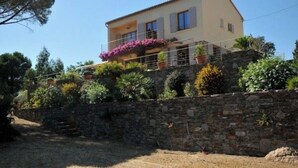 A two-story house with a stone wall in front, surrounded by various plants and trees. - Beautiful villa with sea view, 2.5km from the beach, in the heart of nature   (Bormes-les-Mimosas)