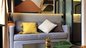 Un salon moderne avec un canapé gris, des coussins jaunes et blancs, une petite table ornée d’un objet décoratif et une grande feuille verte.