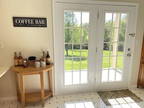 A room with a coffee bar sign, a wooden table with coffee mugs and bottles, and a view of a green lawn through double doors.